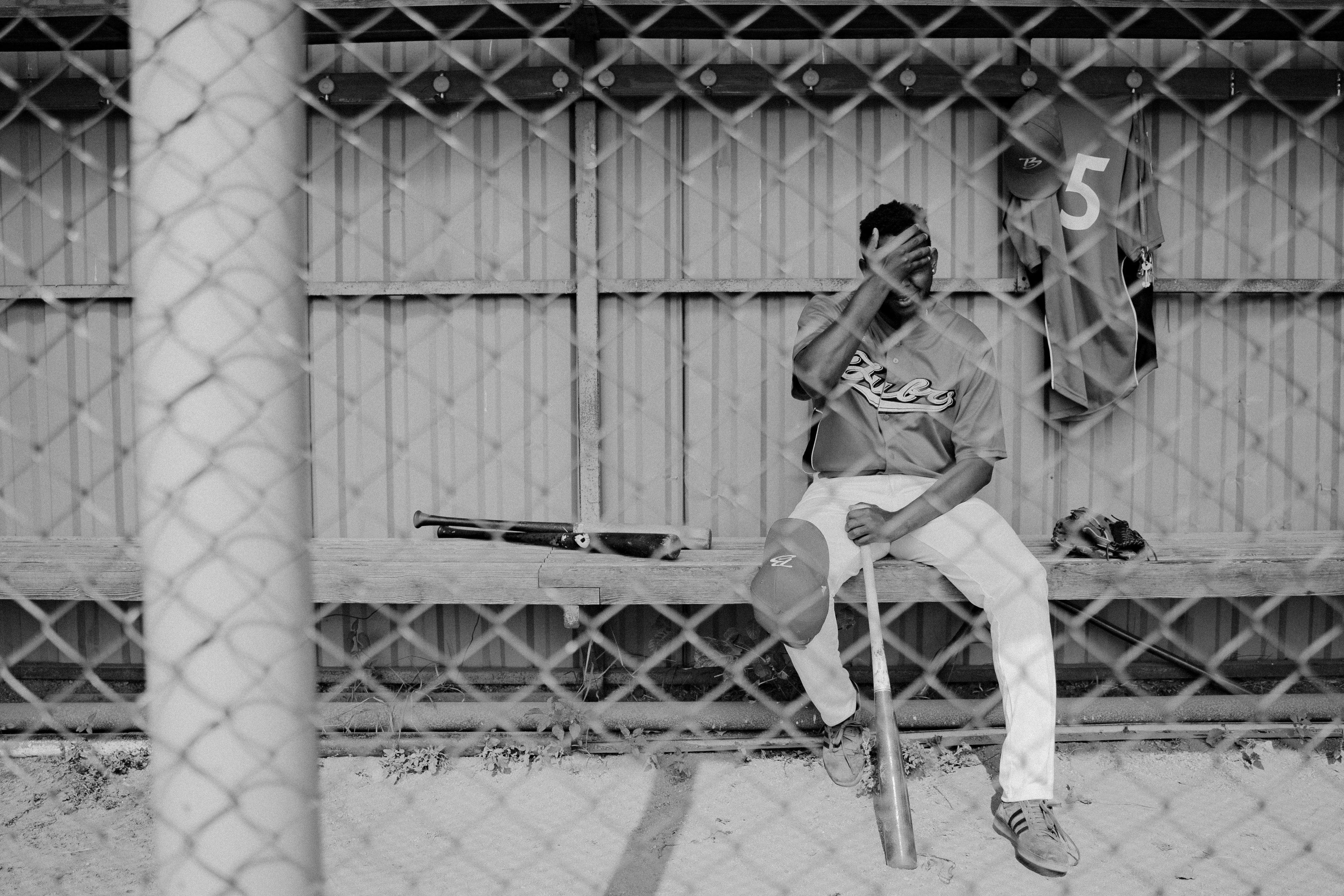 A baseball player sitting in the dugout, holding his head with his right hand. His cap is propped on his right kneecap.