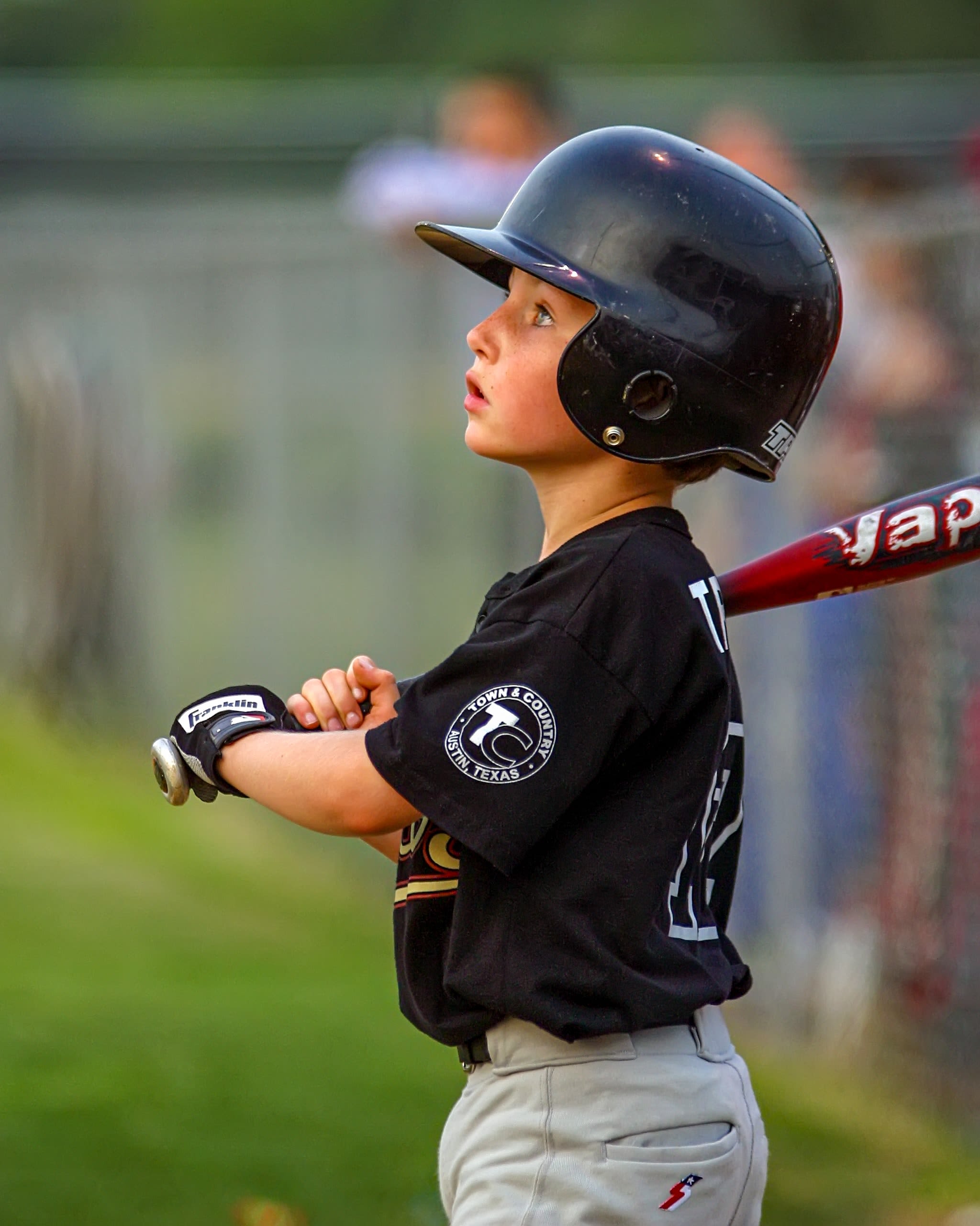 A young child holding a metal baseball bat and wearing a black batting helmet. He is looking upwards and appears to be preparing to swing the bat.