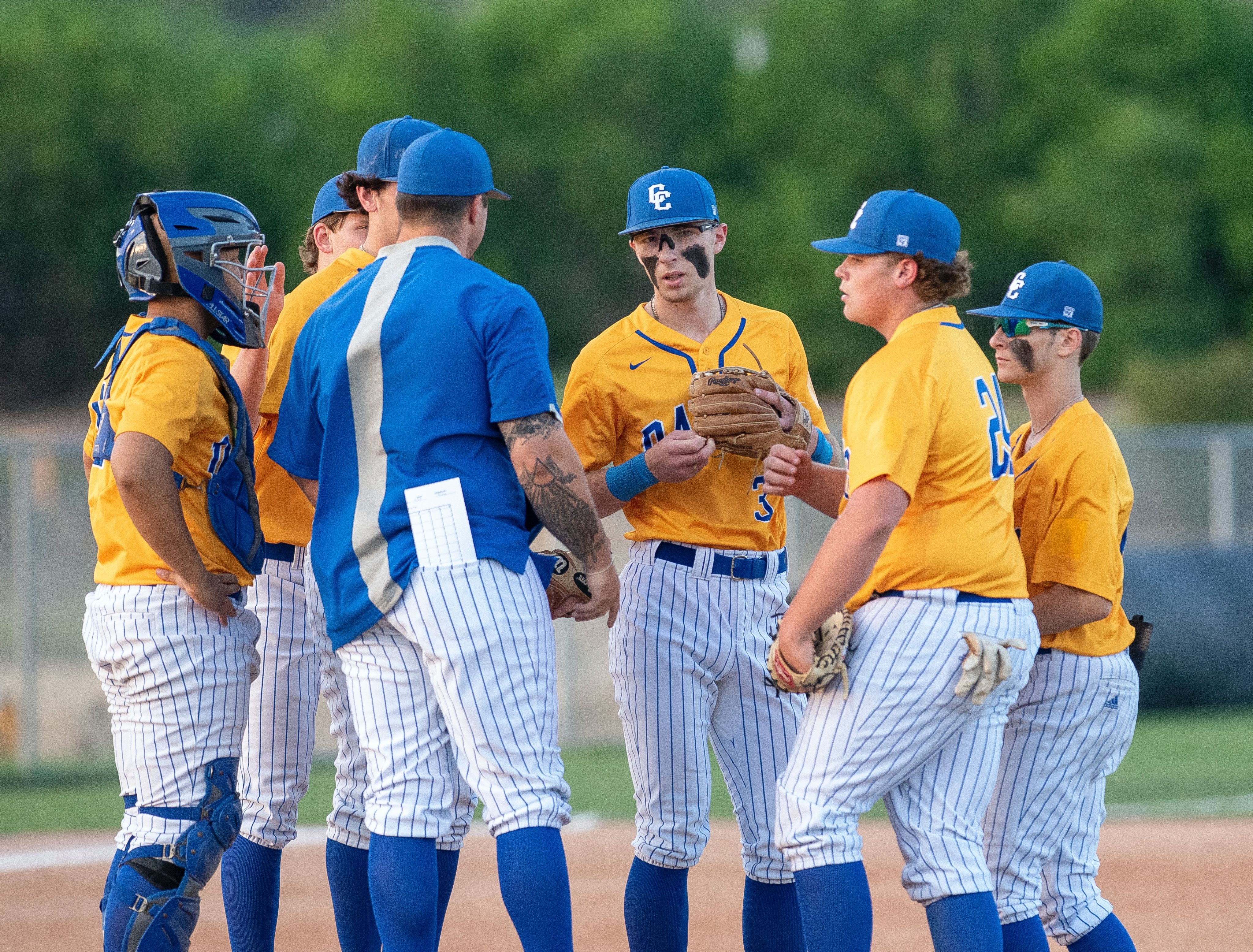 A baseball team wearing yellow jerseys meets in a huddle. A coach, wearing a blue jacket, is having a discussion with his players.